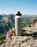 Sage insulated stainless steel water bottle with black threaded lid, on rocky viewpoint