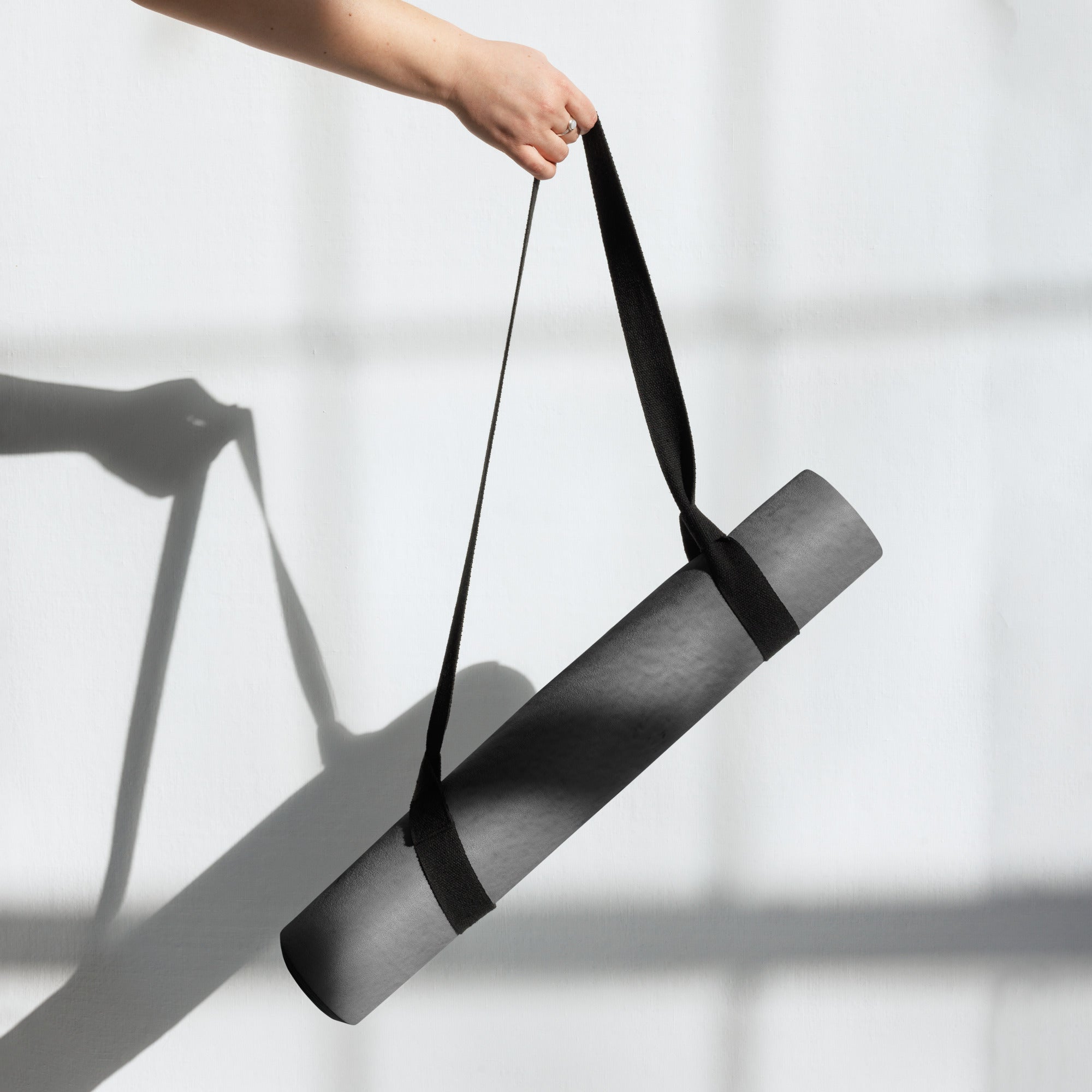 Rolled dark gray yoga mat with black carrying strap held up by hand. Eco Black Dark gray natural rubber yoga mat with Suede top 