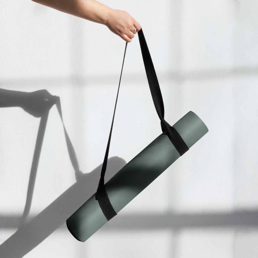 Person carrying a rolled cypress green yoga mat with a black strap.