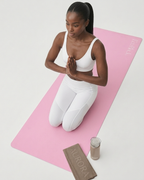A woman in a white sports bra and leggings kneels in a prayer pose on a pink yoga mat. A folded brown towel and a small bottle are placed beside the mat on a clean white studio background.