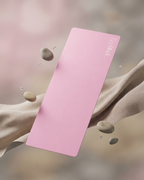 A pink rectangular yoga mat with the brand name 'Yasmani' printed in white near the top right corner, floating diagonally between draped beige fabric and floating smooth stones against a soft, blurred pink background.