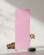 A pink yoga mat standing upright on a light surface, styled with a dried lotus flower, a dark stone, and a piece of cork on a soft neutral background. The brand name 'leo pack.