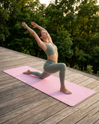 A woman in a sage green activewear set performs a yoga pose on a pink yoga mat placed on a wooden deck with a forest background.