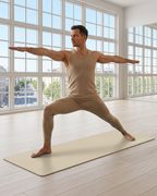 A man in a beige sleeveless tank top and matching leggings performs a yoga pose on a light beige mat in a bright room with large windows and light wood floors.