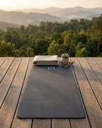 Twilight slate yoga mat laid flat on a wooden deck outdoors, with small folded cloth beside it.