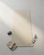 A rectangular beige yoga mat with the brand name 'keshi' printed in elegant script near the top right, styled on a textured gray concrete surface with soft diagonal shadows, a small dark stone, a beige fabric scrap, and a sprig of eucalyptus.