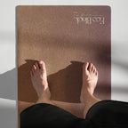 Brown natural rubber yoga mat with feet visible on top; logo centered near top edge.