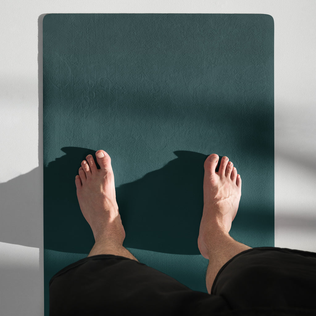 Deep teal yoga mat close-up with textured surface and visible feet resting on it.