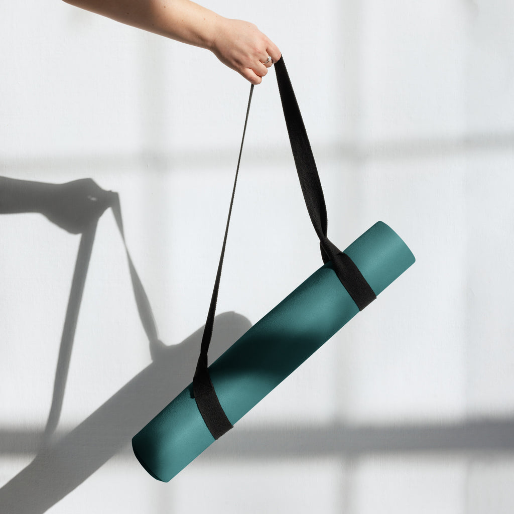 Deep teal natural rubber yoga mat being lifted using a black carrying strap.